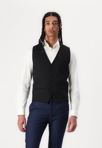 Gilet noir ajusté avec cinq boutons, porté sur une chemise blanche. Un pantalon de costume bleu complète la tenue. Tissu lisse avec une coupe structurée.