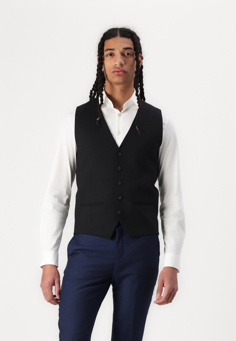 Gilet noir ajusté avec cinq boutons, porté sur une chemise blanche. Un pantalon de costume bleu complète la tenue. Tissu lisse avec une coupe structurée.