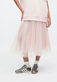 Světlounce růžová tylová sukně s vrstvenými záhyby, doplněná šedobílými teniskami. Oblečení má volný střih, zdůrazňující texturu a průhlednost.