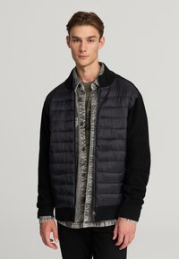 Giacca a vento nera con maniche a coste nere e colletto alto, indossata sopra una camicia a quadri grigia. Presenta una cerniera e dettagli cuciti.