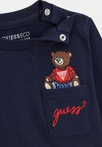 Marineblaues Baumwoll-T-Shirt mit einem gestickten Bären in einem roten Pullover und blauen Hosen, mit einer Tasche, auf der das Wort "guess" in roter Schrift zu sehen ist.