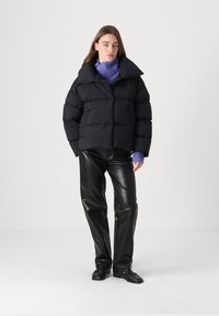 Sort pufferjakke med høj krave, quiltet design og oversized pasform, parret med sorte læderbukser og et lilla strikket tørklæde.