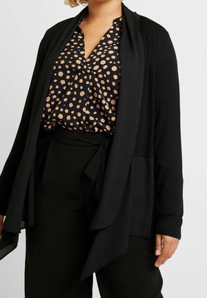 Femme portant un cardigan noir drapé à ouverture avant, un pantalon noir avec taille nouée et un chemisier noir à motif à pois beige.
