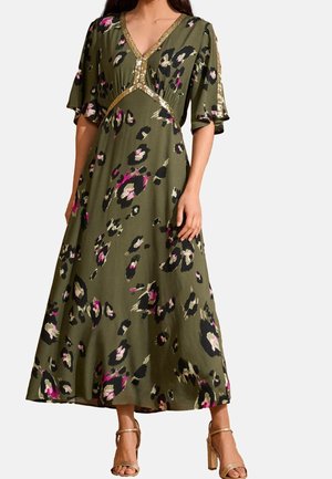 Donna che indossa un vestito lungo verde oliva con motivo floreale nero, rosa e beige e rifinitura in paillettes dorate sullo scollo e sulle maniche.