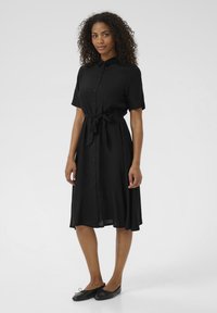 Robe noire à manches courtes, avec un devant boutonné, une taille ceinturée et une jupe fluide. Le tissu lisse et le col ajoutent un détail structuré.