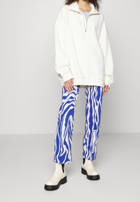 Sweatshirt blanc à fermeture éclair, pantalon à rayures zébrées bleu et blanc, et bottines crème avec semelles noires. Coupe décontractée, texture douce.