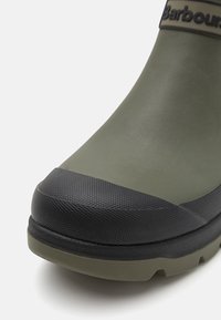 Zöld gumicsizma fekete, textúrázott orrral és talppal, sima testtel, a felső részen a "Barbour" márkanevével.