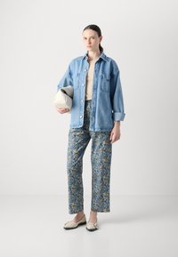 Camicia di denim, top beige, pantaloni blu con motivo floreale e ballerine color crema. La modella tiene una borsa chiara, in piedi contro uno sfondo neutro.