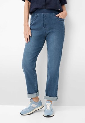 Persoon draagt opgerolde blauwe jeans, lichtblauwe en witte sneakers en een donkerblauw shirt, met één hand in de zak en de andere ontspannen.