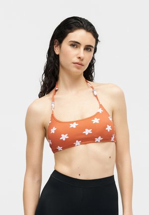 Joven mujer con cabello largo y oscuro que lleva un top de bikini halter naranja con estampado floral blanco y braguitas negras de cintura alta.