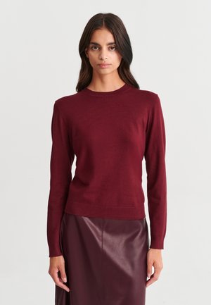 Femme aux longs cheveux foncés portant un pull ajusté bordeaux et une jupe en cuir bordeaux assortie, debout devant un fond uni.