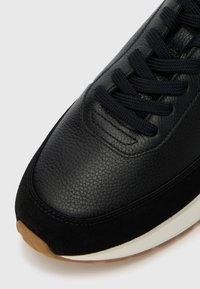 Črne usnjene superge s semiš akcenti. Imajo teksturirano površino, ravne črne vezalke in gumijast podplat s svetlo rjavim dnom.