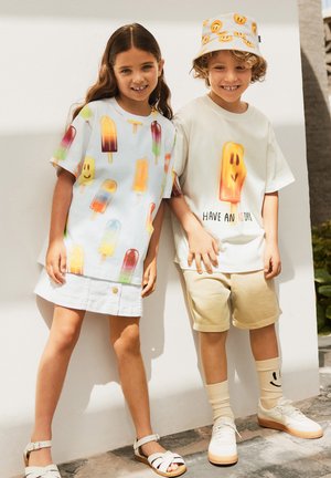 Dos niños posan: uno lleva un vestido blanco con estampados de paletas de colores, el otro lleva una camiseta crema con un gráfico de paleta y pantalones cortos beige.
