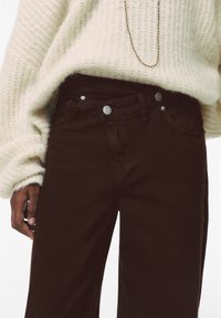 Jeans in denim marrone con vita alta, dotati di hardware argentato, chiusura con bottone e un maglione di colore crema testurizzato indossato sopra.