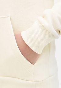 Sweatshirt de couleur crème avec un tissu doux et texturé. Comprend une poche kangourou et des poignets côtelés pour un ajustement confortable.
