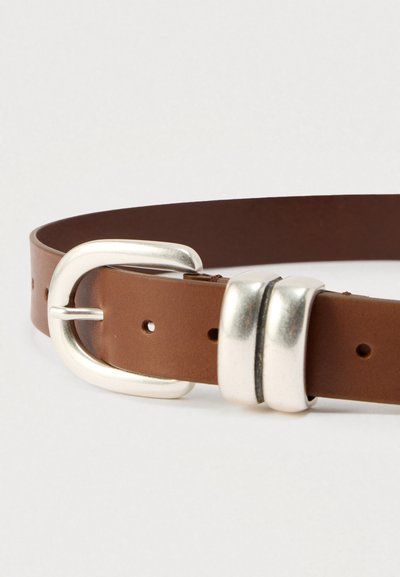 Καφέ δερμάτινη ζώνη με ασημένια μεταλλική αγκράφα. Διαθέτει λεία υφή, στρογγυλεμένο σχήμα και πολλαπλές τρύπες ρύθμισης κατά μήκος της ζώνης.