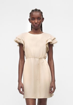 Kobieta w beżowej sukience do kolan z marszczonymi krótkimi rękawami i cienkim paskiem w talii, stojąca na jednolitym białym tle.
