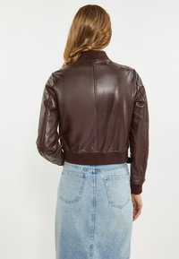 Bruin leren bomberjack met ribbed manchetten en kraag. Gevuld over een lichtblauwe denimrok met twee achterzakken. Gladde textuur.