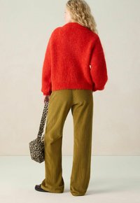 Frau mit lockigem blonden Haar, die einen roten, flauschigen Pullover und olivegrüne, weite Cordhosen trägt, während sie eine Tasche im Leopardenmuster hält und von hinten schaut.