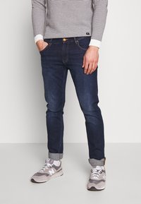 Mörkblå denimjeans med slim fit, som har en klassisk femficksdesign, orange sömmar och uppvikta nederkanter. I kombination med grå sneakers.