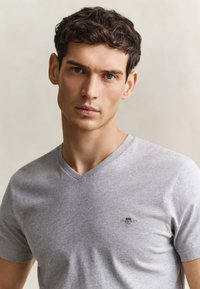 Ung mand med kort mørkt hår iført en lysegrå T-shirt med V-udskæring og en lille broderet logo på brystet mod en ensfarvet baggrund.