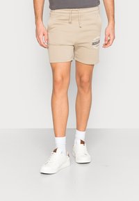 Beige bavlněné šortky s elastickým pasem a šňůrkou, s potiskem loga. V kombinaci s bílými teniskami a ponožkami do poloviny lýtek.