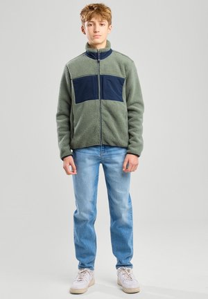Giacca in pile verde tenue con dettagli blu navy. Presenta un colletto alto, zip a piena lunghezza e due tasche frontali. Abbinata a jeans azzurri chiari e sneaker bianche.