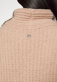Beige gebreide trui met hoge kraag, voorzien van een gestructureerd patroon en een klein metalen logootje op de achterkant.