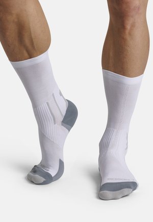 Gambe con calzini bianchi a costine con talloni e punte grigi, un piede appoggiato e l'altro leggermente sollevato sulle punte, su sfondo bianco.