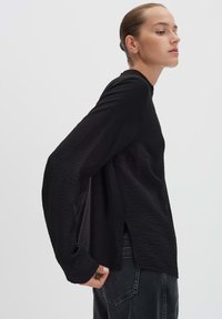 Donna in un top a maniche lunghe nera e testurizzata e jeans scuri, in piedi di lato contro uno sfondo grigio chiaro semplice.