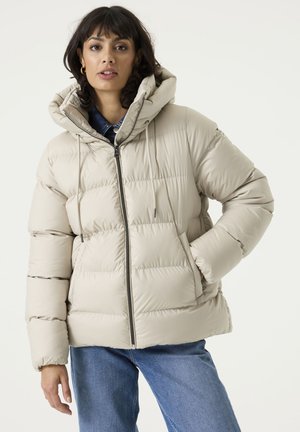 Frau trägt eine hellbeige Steppjacke mit Kapuze, blaue Jeans und goldene Creolen und steht vor einem schlichten, hellen Hintergrund.