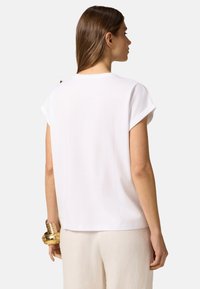 Camiseta blanca de manga corta con un corte relajado. Textura suave, hombros ligeramente caídos y un dobladillo curvado, combinada con pantalones beige claro.