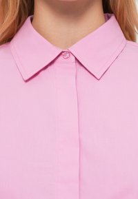 Ροζ μπλούζα με γιακά και κούμπωμα με κουμπιά στο λαιμό. Το ύφασμα φαίνεται λείο και ελαφρύ, με καθαρό φινίρισμα.