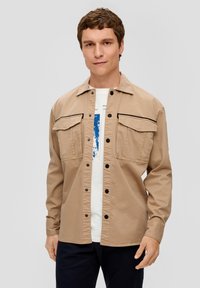 Giacca button-up color cammello con due tasche sul petto e dettagli neri. Realizzata in un tessuto morbido, presenta maniche lunghe e una vestibilità comoda.