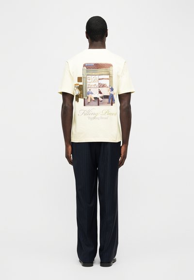 Άνδρας που φοράει κρεμ μπλουζάκι με εικονογράφηση καταστήματος αρτοποιείου και κείμενο "Filling Pieces Breaking Bread" στην πλάτη, σε συνδυασμό με σκούρο παντελόνι με λεπτές ρίγες.