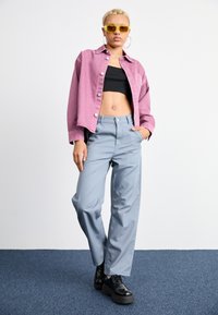 Veste en denim rose pâle, crop, haut noir ajusté, pantalon large gris clair et chaussures à plateforme noires épaisses. Lunettes de soleil jaunes vives.