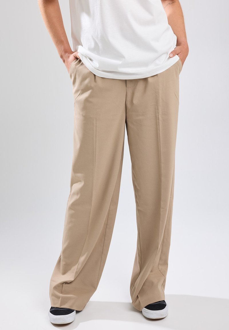Beige wide-leg trousers with a smooth texture, front pleats, and side pockets, paired with black and white sneakers and a white t-shirt.