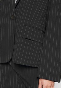Veste de costume noire à fines rayures avec bouton et poche à rabat, faisant partie d’un pantalon assorti, portée par une personne avec une main visible sur le côté.