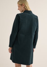 Frau mit blonden Haaren, die ein dunkelgrünes langärmliges Kordkleid trägt, mit dem Rücken zur Kamera, Augen geschlossen und einem leichten Lächeln.