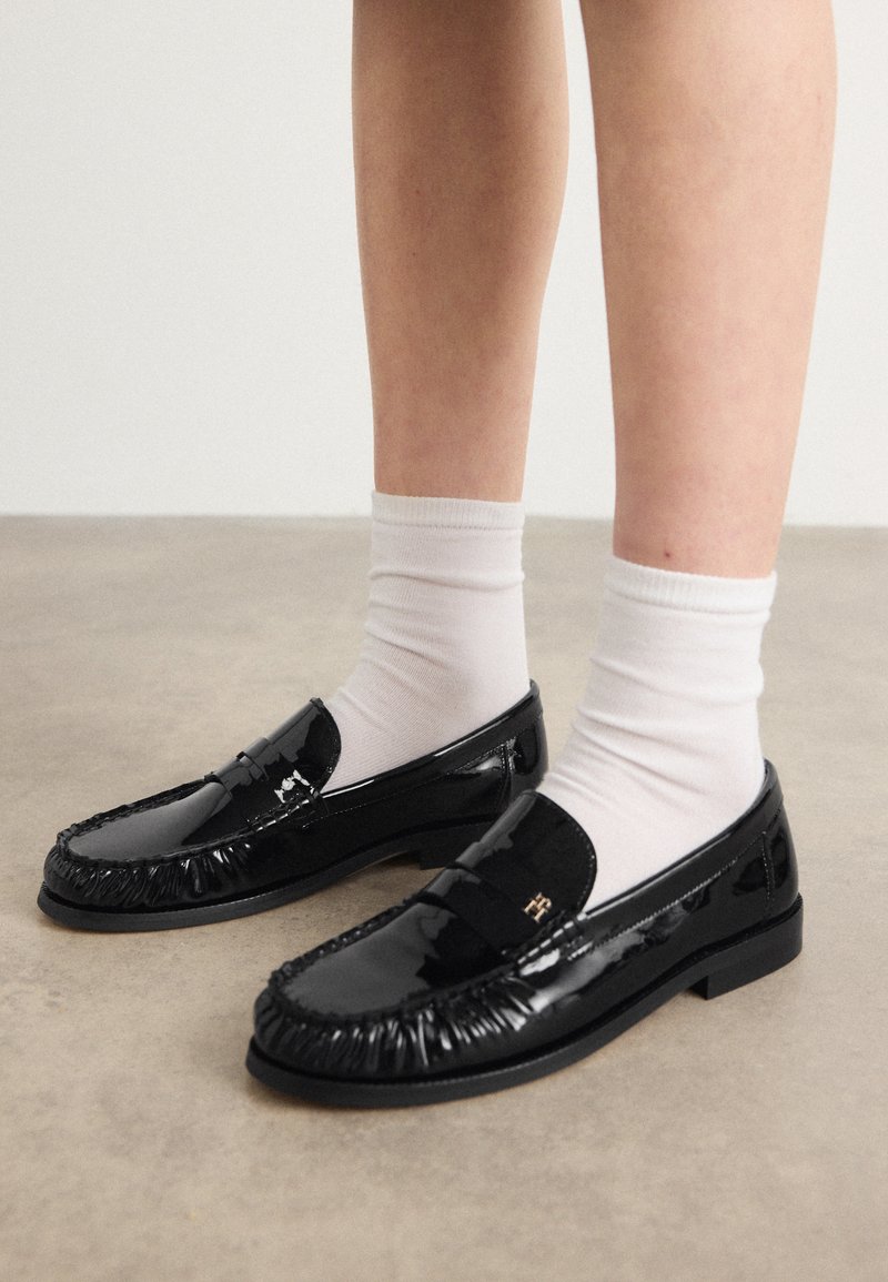 Schwarze Lacklederloafer mit fransigem Design, akzentuiert durch ein kleines Metallsymbol und getragen mit weißen Knöchelsocken. Flache Sohle.