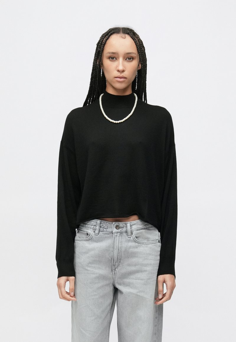 Maglione nero corto con maniche lunghe e collo alto, abbinato a jeans grigio chiaro a vita alta. La modella indossa una collana di perle.