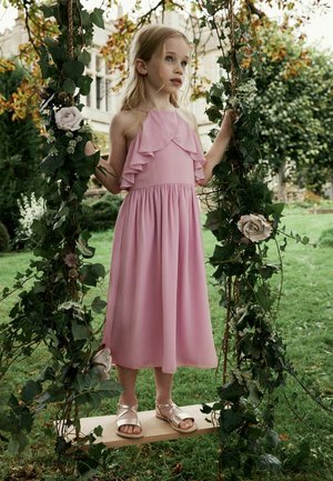 Young girl in pink dress standing on a flower-adorned wooden swing in a green garden with trees and a building in the background.