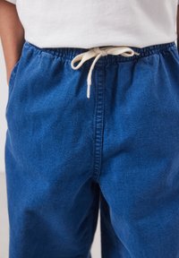 Personne portant un pantalon à taille élastique bleu avec un cordon blanc noué en nœud papillon et une chemise blanche rentrée.