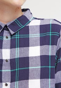 Navy blue and white plaid shirt with green accents, featuring a collared neck, button-front closure, and herringbone texture.