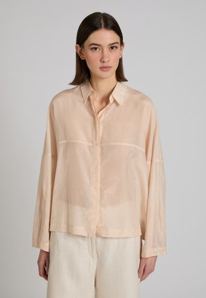 Donna che indossa una camicetta lunga a maniche lunghe beige, leggera e trasparente, e pantaloni chiari, in piedi contro uno sfondo grigio uniforme.