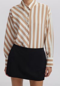 Camicia button-up con strisce verticali beige e bianche, maniche lunghe e colletto classico; abbinata a una gonna nera aderente.
