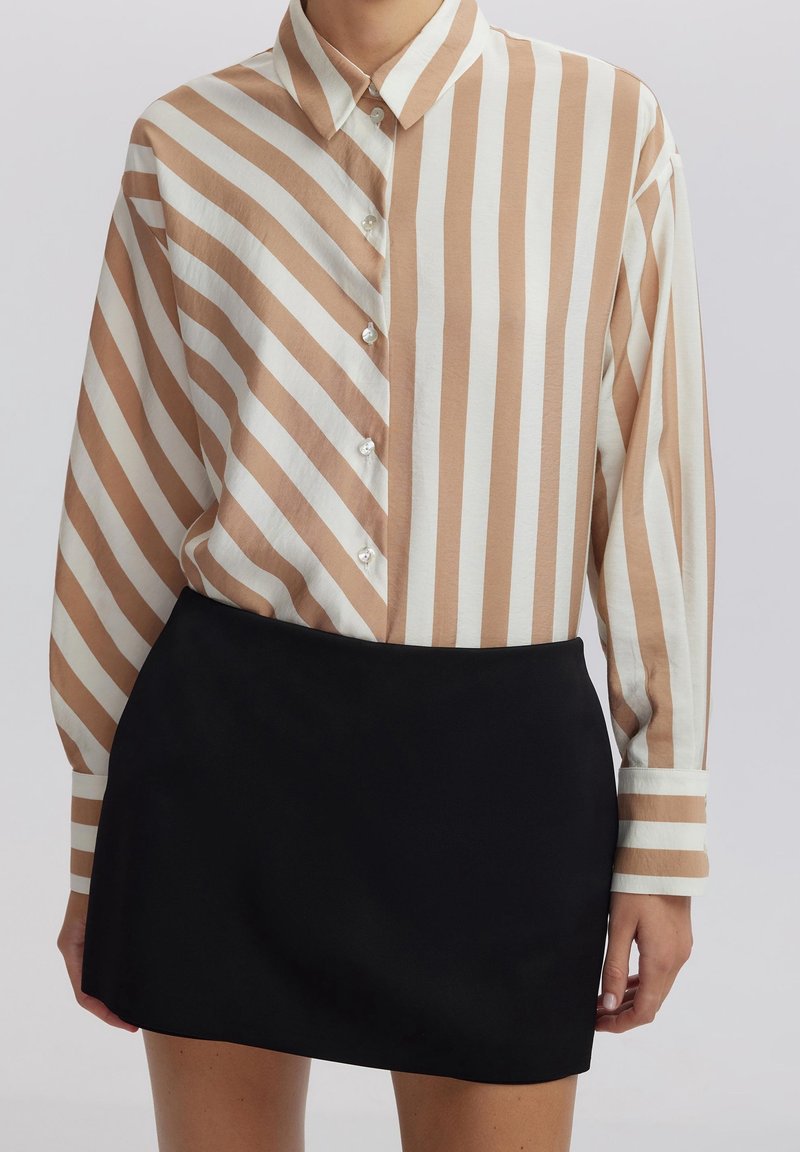 Camicia button-up con strisce verticali beige e bianche, maniche lunghe e colletto classico; abbinata a una gonna nera aderente.