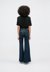 Göndör hajú személy fekete pólóban és magas derekú, széles szárú kék farmert viselve, háttal áll fehér háttér előtt.