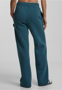Pantalones de chándal en color verde azulado con un ajuste relajado, cinturilla elástica, bolsillos laterales y una textura suave. Detalles de costura visibles a lo largo de las piernas.