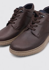 Chaussures en cuir marron à lacets avec des semelles texturées beige, des bouts arrondis et des œillets en métal, photographiées sur un fond clair.